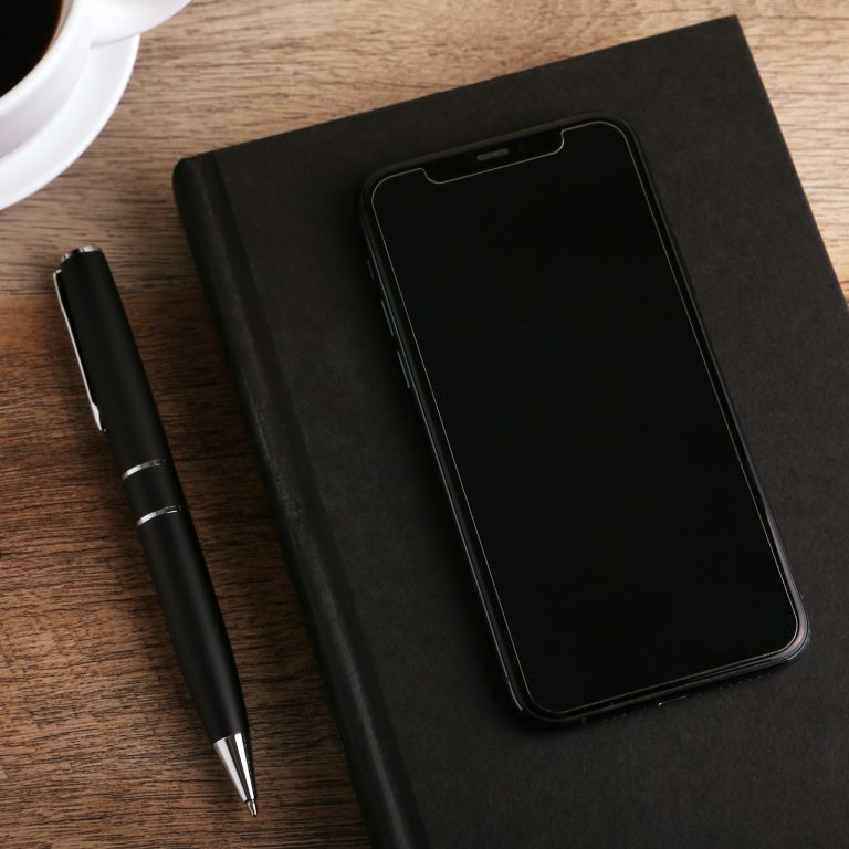 Black smartphone on a closed notebook beside a pen and coffee cup.
