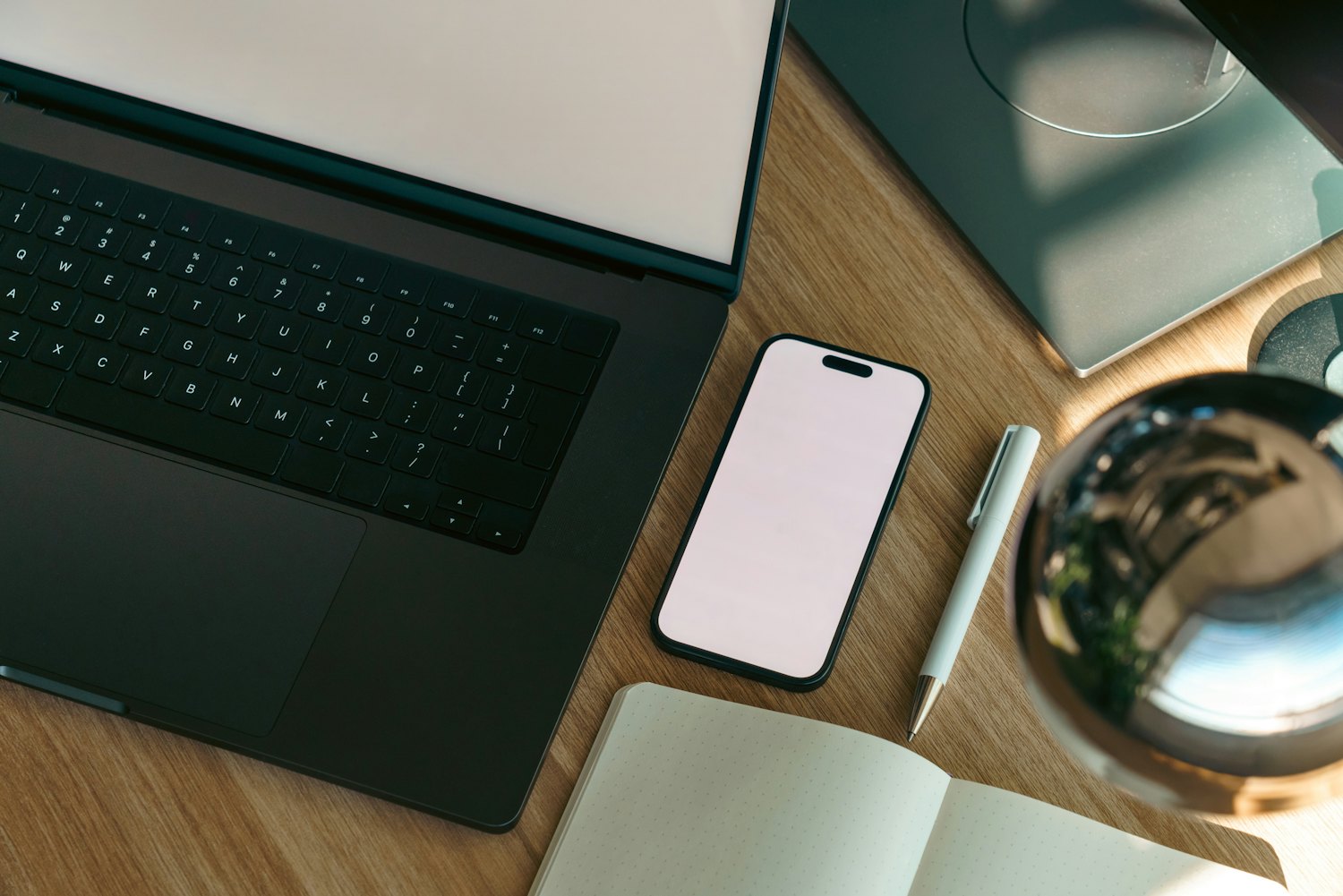 Blank smartphone and laptop screens beside an open notebook and pen on a wooden desk.